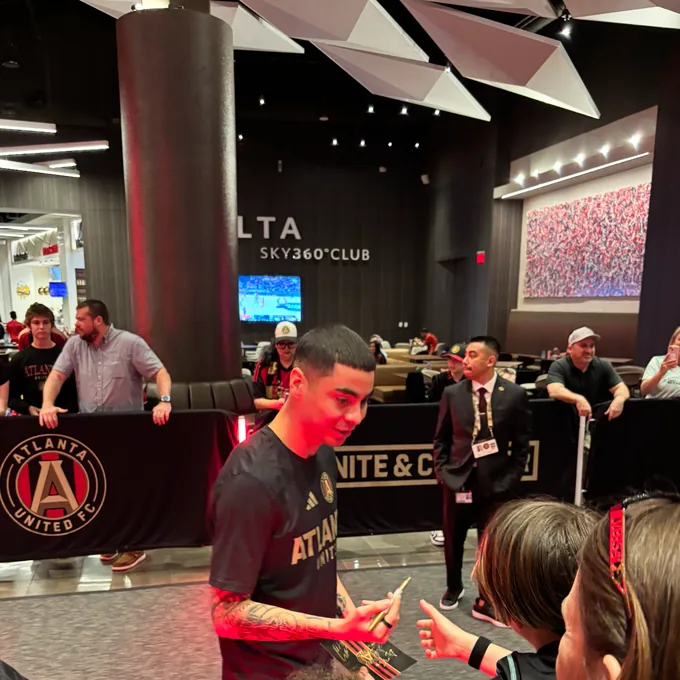 Miguel Almirón, in an Atlanta United training top, signing a card for a young fan in the stadium club.