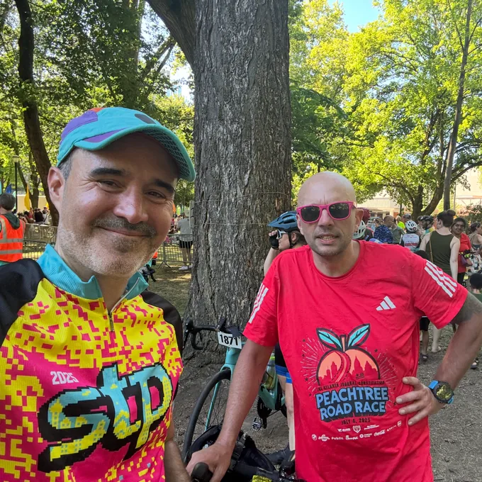 Thomas in an STP cycling jersey beside his bike, with a friend in a Peachtree Road Race shirt, under trees at a rest stop.